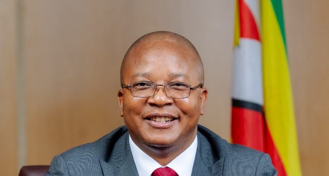 Zimbabwe Mines Minister Polite Kambamura seated at a desk with the national flag behind him.