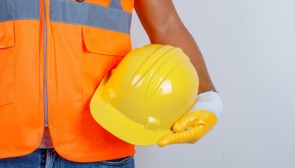 Person in orange safety vest holding yellow hard hat against plain background.