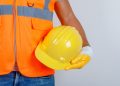 Person in orange safety vest holding yellow hard hat against plain background.