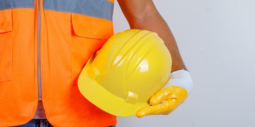 Person in orange safety vest holding yellow hard hat against plain background.