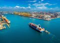 Aerial view of a busy commercial port with container ships, cranes, and stacked cargo containers.