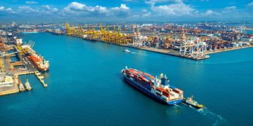 Aerial view of a busy commercial port with container ships, cranes, and stacked cargo containers.