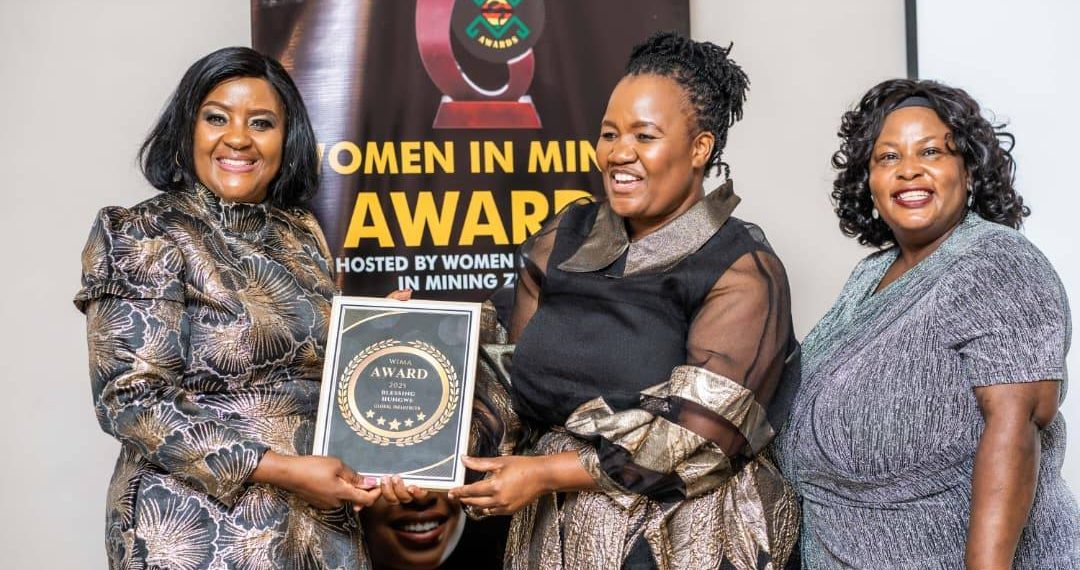 Two women hold an award plaque during the Women in Mining Service Excellence Awards in Harare, with a banner in the background.