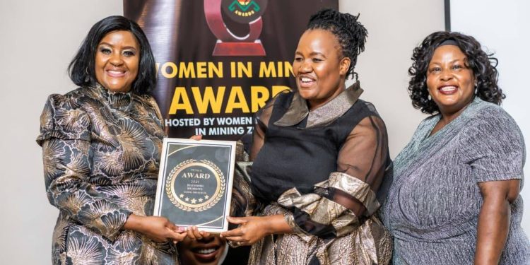 Two women hold an award plaque during the Women in Mining Service Excellence Awards in Harare, with a banner in the background.
