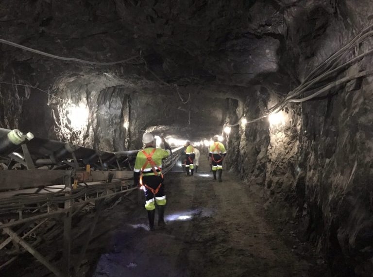 Underground mining tunnel at Blanket Mine in Zimbabwe, with workers in safety gear walking past conveyor systems.