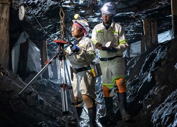 Two underground mine workers using surveying equipment in a dimly lit tunnel.