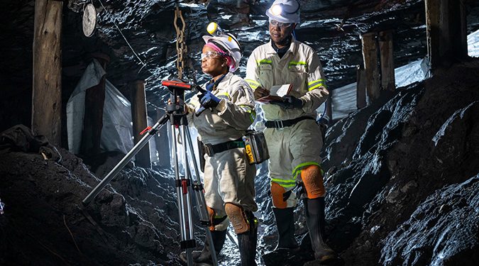 Two underground mine workers using surveying equipment in a dimly lit tunnel.