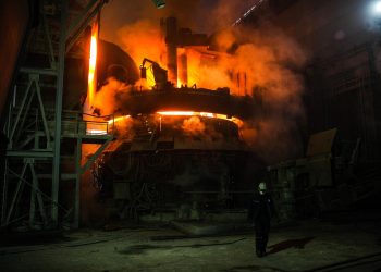 Worker in protective gear walking inside a metal smelting plant with a bright furnace in the background.