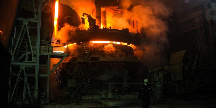 Worker in protective gear walking inside a metal smelting plant with a bright furnace in the background.