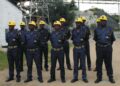 Mine security guards in blue uniforms and yellow helmets stand in formation at an industrial site.