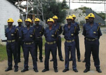 Mine security guards in blue uniforms and yellow helmets stand in formation at an industrial site.
