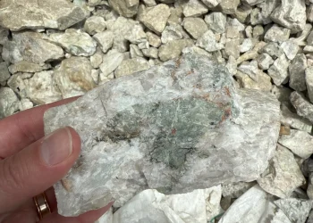 Close-up of a hand holding a white and green lithium-bearing rock sample over crushed stones at the Fonlo Project in Nigeria.