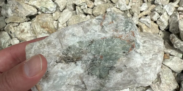 Close-up of a hand holding a white and green lithium-bearing rock sample over crushed stones at the Fonlo Project in Nigeria.
