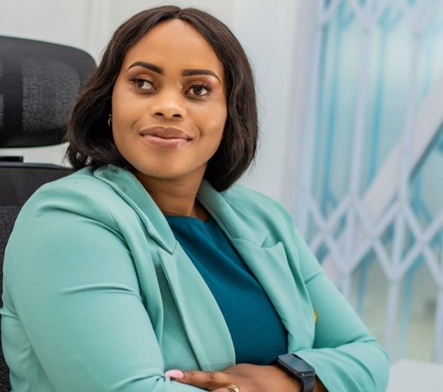 A woman in a teal blazer sits on a dark office chair indoors, wearing a smartwatch, with a window and light wall in the background.