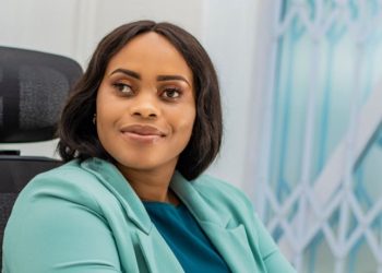 A woman in a teal blazer sits on a dark office chair indoors, wearing a smartwatch, with a window and light wall in the background.