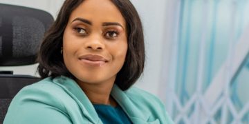 A woman in a teal blazer sits on a dark office chair indoors, wearing a smartwatch, with a window and light wall in the background.