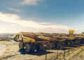 A row of large haul trucks parked at an open-pit mine, with additional mining vehicles and earthworks visible in the background.