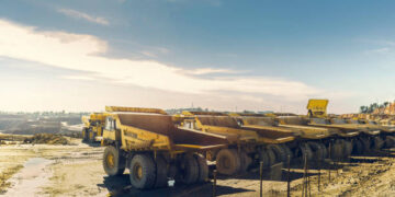 A row of large haul trucks parked at an open-pit mine, with additional mining vehicles and earthworks visible in the background.