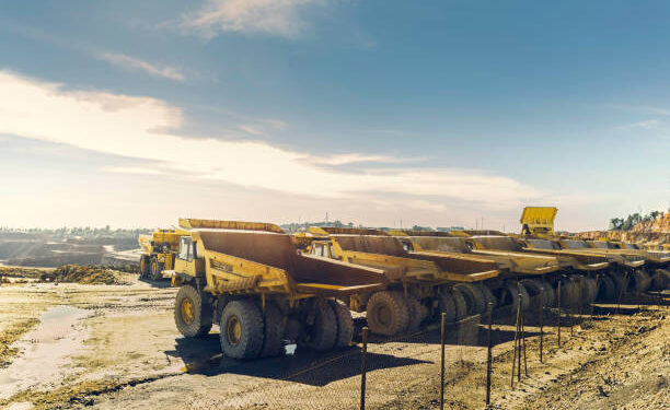 A row of large haul trucks parked at an open-pit mine, with additional mining vehicles and earthworks visible in the background.