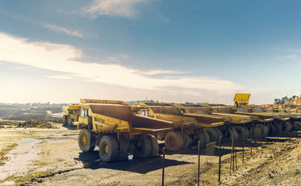 A row of large haul trucks parked at an open-pit mine, with additional mining vehicles and earthworks visible in the background.