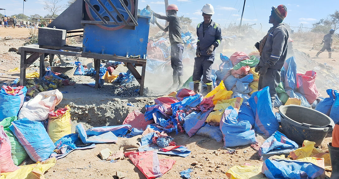 Three miners in hard hats operate a rock-crushing mill at a small-scale gold mining site, with dust rising and sacks of crushed ore piled in the foreground.