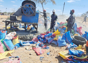 Three miners in hard hats operate a rock-crushing mill at a small-scale gold mining site, with dust rising and sacks of crushed ore piled in the foreground.