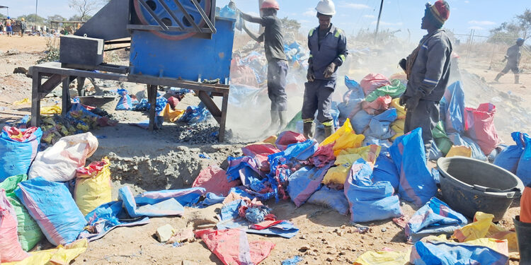 Three miners in hard hats operate a rock-crushing mill at a small-scale gold mining site, with dust rising and sacks of crushed ore piled in the foreground.