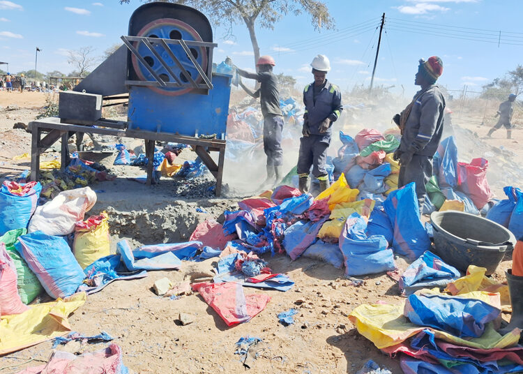 Three miners in hard hats operate a rock-crushing mill at a small-scale gold mining site, with dust rising and sacks of crushed ore piled in the foreground.