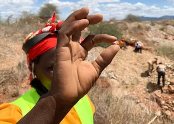 A woman miner holds a small mineral sample while other miners work at an artisanal mining site