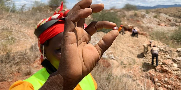 A woman miner holds a small mineral sample while other miners work at an artisanal mining site