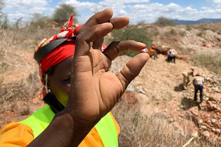 A woman miner holds a small mineral sample while other miners work at an artisanal mining site