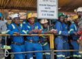 Miners in blue overalls and hard hats stand behind a railing beside a sign reading How Mine Main Shaft, Safe Work Load 2.6 tons