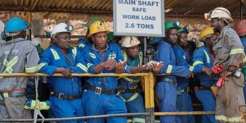 Miners in blue overalls and hard hats stand behind a railing beside a sign reading How Mine Main Shaft, Safe Work Load 2.6 tons