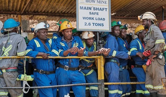Miners in blue overalls and hard hats stand behind a railing beside a sign reading How Mine Main Shaft, Safe Work Load 2.6 tons