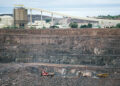 Haul trucks and an excavator work in the open pit at Orapa diamond mine in Botswana, with a processing plant and conveyor above