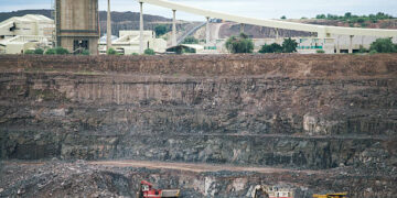 Haul trucks and an excavator work in the open pit at Orapa diamond mine in Botswana, with a processing plant and conveyor above