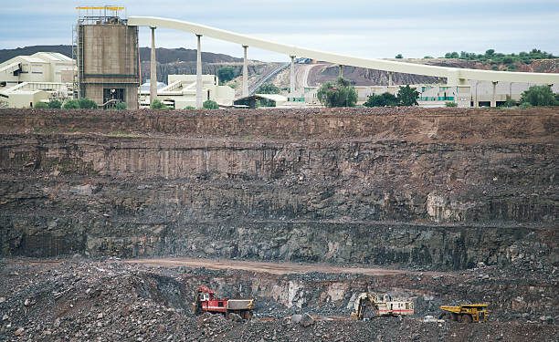 Haul trucks and an excavator work in the open pit at Orapa diamond mine in Botswana, with a processing plant and conveyor above