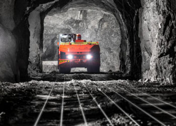 Underground mining truck travelling through a mine tunnel illuminated by vehicle headlights.
