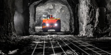Underground mining truck travelling through a mine tunnel illuminated by vehicle headlights.