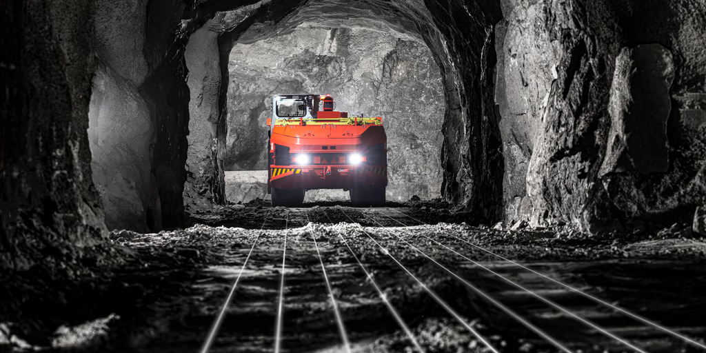 Underground mining truck travelling through a mine tunnel illuminated by vehicle headlights.
